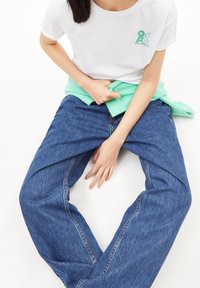 Camiseta blanca de algodón con logo verde, combinada con unos vaqueros de mezclilla holgados en azul. Un suéter de punto verde claro está atado a la cintura.