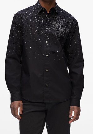 Mężczyzna ubrany w czarną koszulę zapinaną na guziki z rozproszonymi małymi białymi kropkami skoncentrowanymi wokół lewej części klatki piersiowej, tworzącymi subtelny wzór przypominający logo.