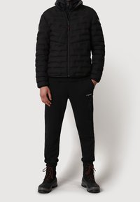Chaqueta acolchada negra con cierre de cremallera, combinada con pantalones de chándal negros y botas negras robustas con cordones rojos, creando un aspecto unificado.