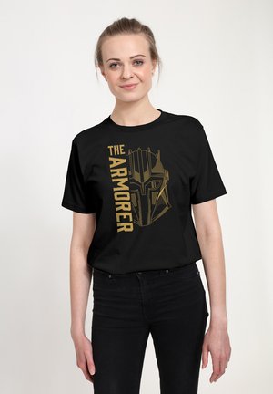 Kobieta nosząca czarny t-shirt z złotym nadrukiem stylizowanego hełmu zbrojnego i pionowym napisem "THE ARMORER" na przodzie.