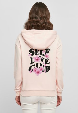 Lys rosa hettegenser med en svart grafisk trykk som leser "SELF LOVE CLUB" dekorert med rosa blomsterdetaljer. Mykt stoff, uformell design.
