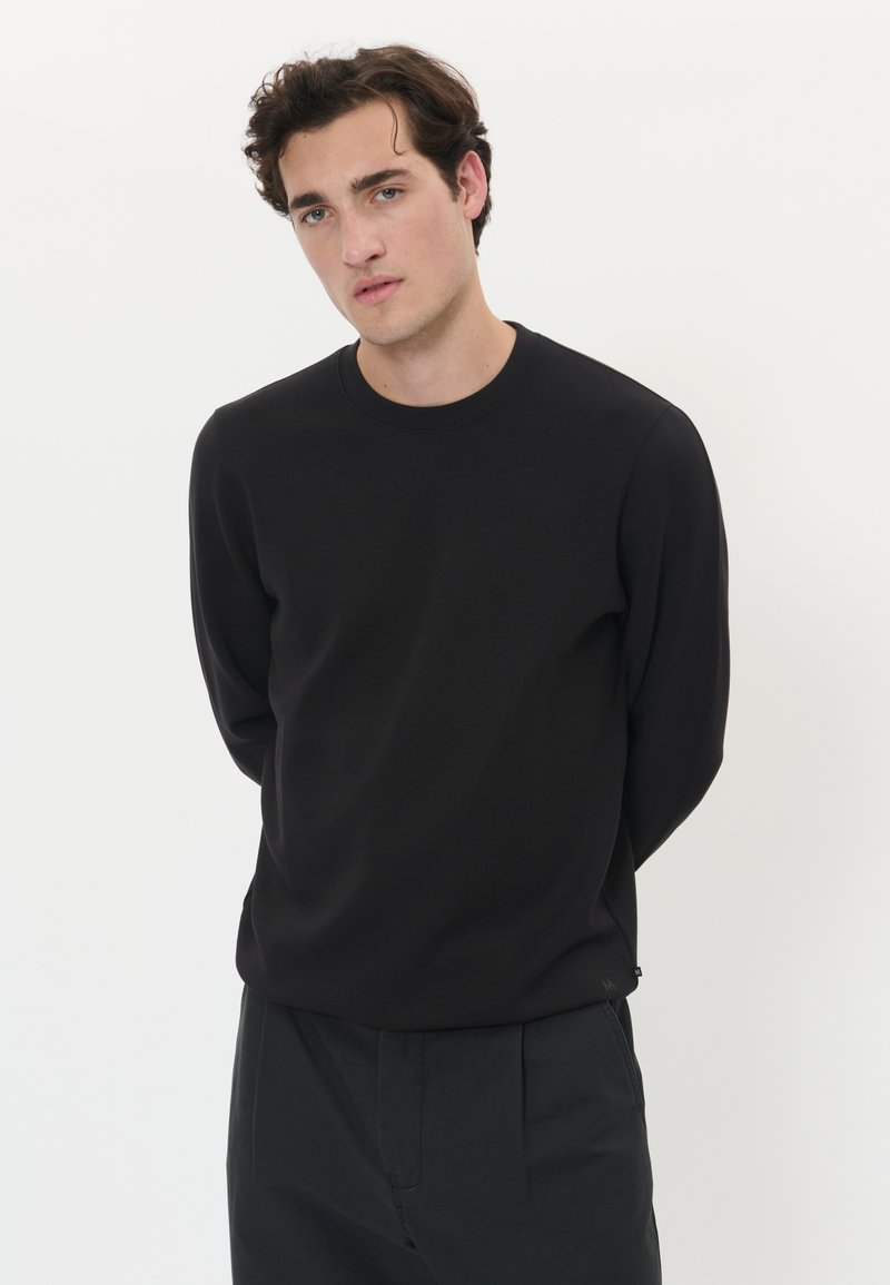 Mladić s tamnom kovrčavom kosom, obučen u običnu crnu dugih rukava majicu i crne hlače, stoji nasuprot obične bijele pozadine.