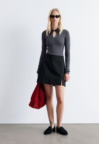 Grauer gerippter Pullover mit Schlitzkragen, schwarzer Minirock mit seitlichem Schlitz, schwarze Slipper und eine rote Wildleder-Tasche.