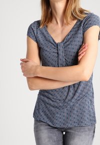 Modrá krátká halenka s geometrickým vzorem, s nařaseným výstřihem a splývavou látkou, v kombinaci se šedými slim fit džíny.