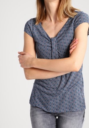 Top blu corto a maniche corte con motivo geometrico, scollatura arricciata e tessuto drappeggiato, abbinato a jeans grigi aderenti.