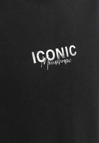 Camiseta de algodón negra con texto impreso en blanco que dice "ICÓNICO" y "Monocromo". Corte ajustado con una textura suave.