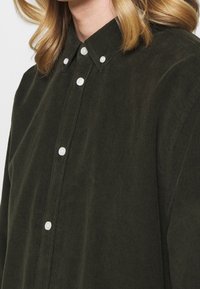 Nærbillede af en lysblond person iført en mørkegrøn fløjlsskjorte med hvide knapper og krave der kan knappes ned.