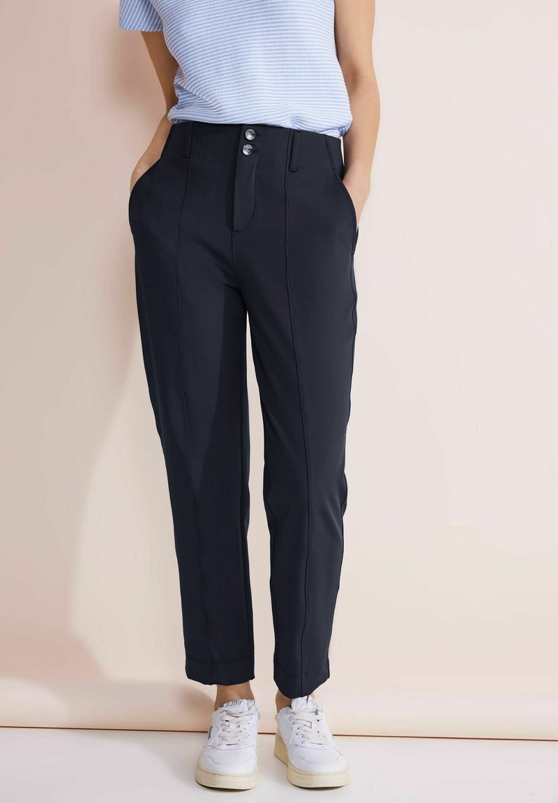 Mörkblå skräddarsydda byxor med knäppt fram, två sidfickor och en avsmalnande passform. Bärs med vita sneakers på en neutral bakgrund.
