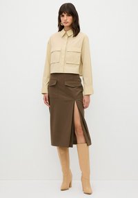 Camicia beige corta con due tasche sul petto, abbinata a una gonna midi marrone con uno spacco laterale e indossata con stivali alti fino al ginocchio beige con tacco.