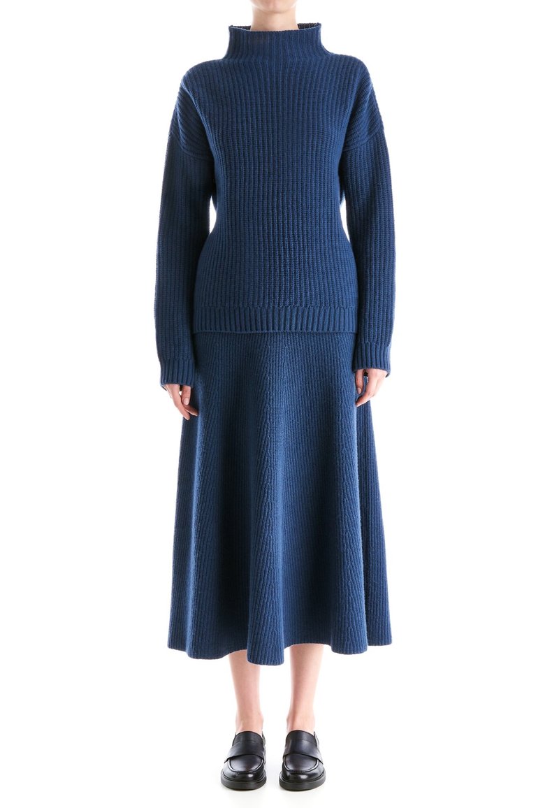 Femme portant un pull en maille côtelée bleu marine à col montant et une jupe midi assortie, associée à des mocassins noirs, debout devant un fond blanc.