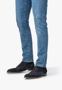 Uomo che indossa jeans slim-fit blu e scarpe da cerimonia in suede blu scuro con lacci, in piedi su una superficie bianca.