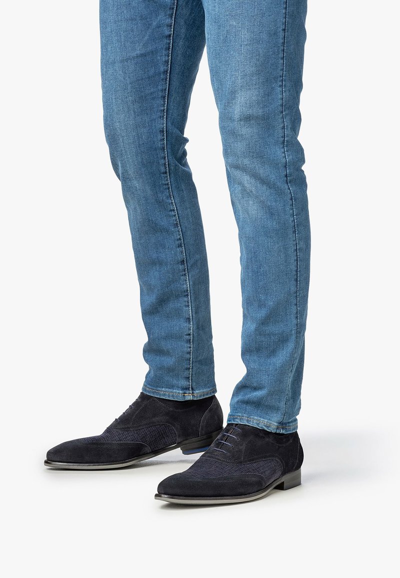 Uomo che indossa jeans slim-fit blu e scarpe da cerimonia in suede blu scuro con lacci, in piedi su una superficie bianca.