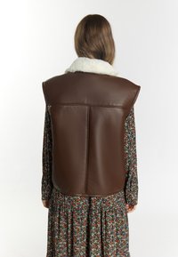 Bruine leren vest met een witte faux bont kraag, ontworpen met een soepele textuur en een achternaaddetail, gedragen over een jurk met bloemenpatroon.