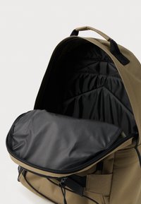 Sac à dos kaki ouvert montrant un compartiment intérieur rembourré noir et une rabat zippé avec des détails sur la poignée et la poche avant.