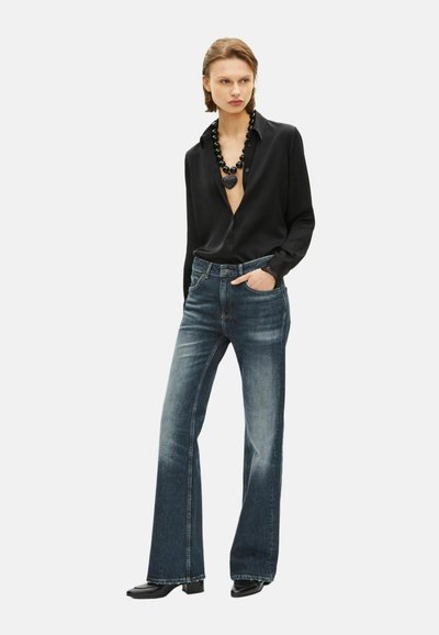 Blusa negra de botones con un escote profundo, combinada con jeans de campana azul oscuro. La modelo lleva un collar negro grueso y botas de tacón.