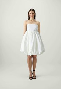 Balta bezplecu kleita ar teksturētu, pūkainu svārku, savilktu viduklī. Modelis nēsā melnas sandales ar siksniņām, ar minimālu dizainu.