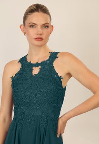 Teal jurk met ingewikkelde bloemenkant overlay, halterhalslijn, sleutelgat detail en vloeiende stof. Opvallende, zachte textuur.