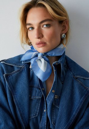 Donna bionda con occhi azzurri che indossa grandi orecchini d'argento, una sciarpa di seta blu e bianca annodata al collo e una giacca di jeans blu scuro.