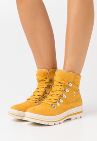 Bottines en daim jaune avec œillets métalliques, semelle épaisse en caoutchouc blanc et col texturé. Lacets assurant un maintien sûr ; aucun motif visible.