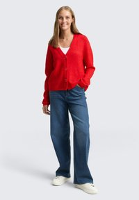 Czerwony, dziergany sweter na guziki z dekoltem w serek, z czterema guzikami z przodu. Nosi się go na białą bluzkę i łączy z niebieskimi spodniami o szerokich nogawkach oraz białymi tenisówkami.