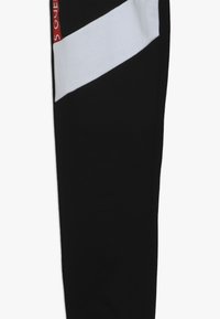 Černé sportovní kalhoty s bílým diagonálním pruhem na boku a červeným logem. Vyrobeny z hladkého, pružného materiálu.