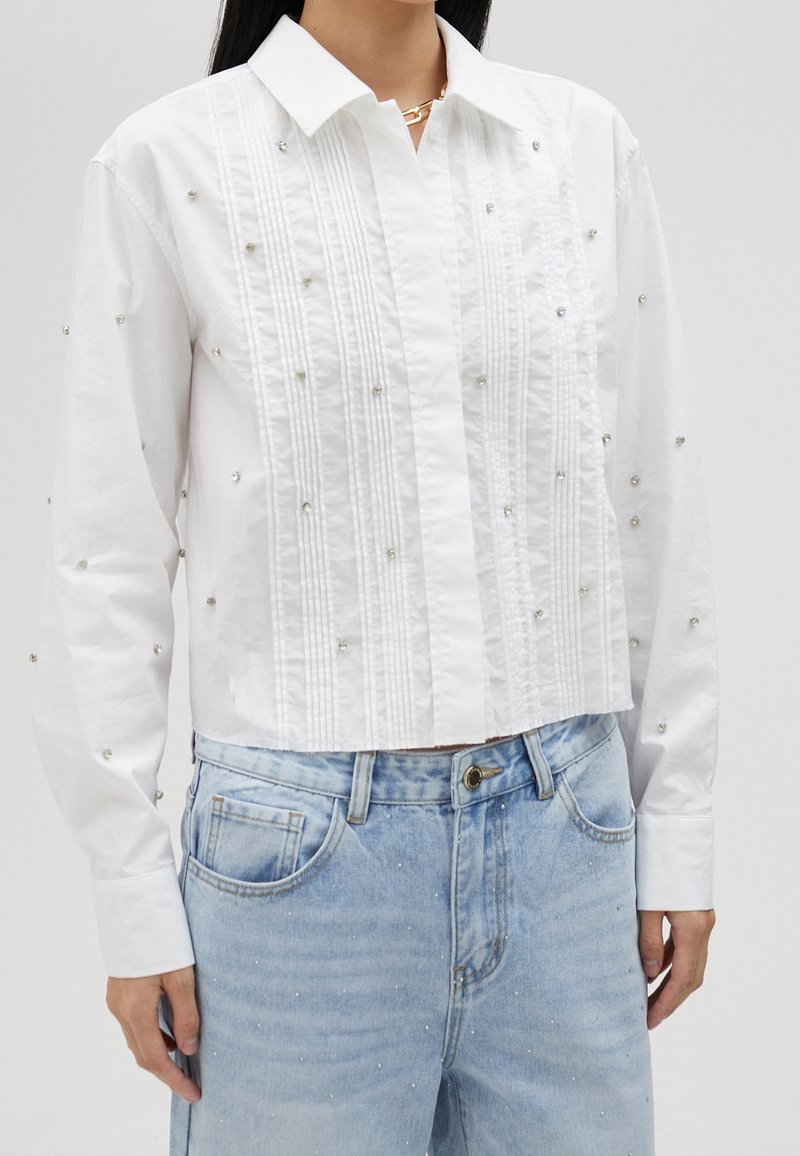 Witte blouse met verticale plooien en verspreide rhinestones. Draag het met lichtblauwe denim jeans met versierde details.
