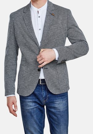 Homme portant un blazer gris à motifs sur une chemise blanche à boutons et un jean bleu, boutonnant le blazer avec sa main droite.