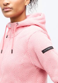 Κοντινό πλάνο ενός ατόμου που φοράει φούτερ με κουκούλα ροζ και λευκό σε σχέδιο ρόμβων, με μαύρο λογότυπο Icepeak στο μανίκι.