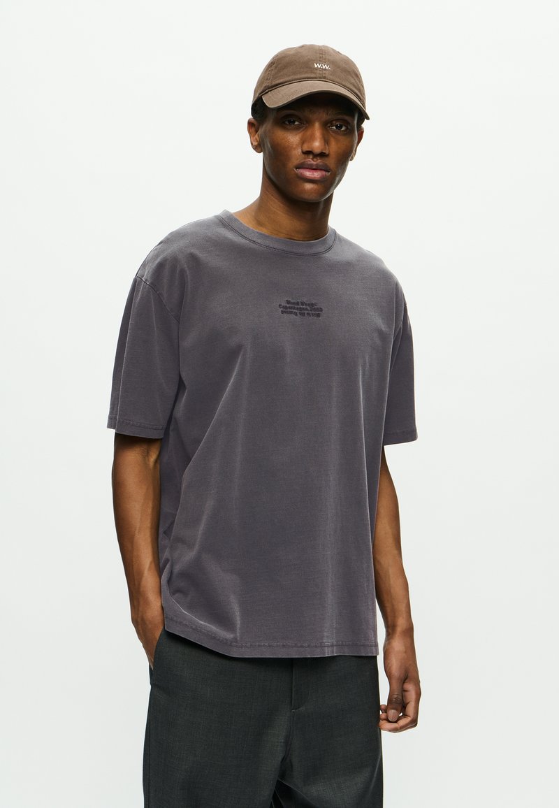 Bărbat îmbrăcat cu un tricou oversized purpuriu estompat, cu un text mic centrat, pantaloni gri închis și o șapcă de baseball maro, pe un fundal alb simplu.