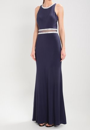 Vrouw in een mouwloze donkerblauwe avondjurk met zilveren bies langs de halslijn en taille, staand tegen een effen achtergrond.