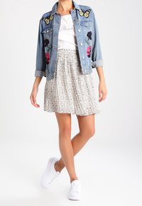 Giacca di jeans con ricami floreali e applicazioni a forma di farfalla, t-shirt bianca, gonna plissettata floreale e sneakers bianche. Outfit casual.