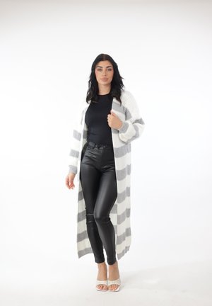 Kvinde iført en lang hvid og gråstribet cardigan, sort top, sorte læderbukser og hvide åbne hæle stående på en hvid baggrund.