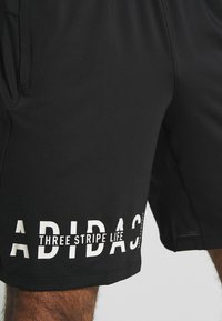 Mustat urheilushortsit, jotka on valmistettu sileästä kankaasta; sivussa olevassa tekstissä lukee vahvalla valkoisella kirjoituksella "ADIDAS THREE STRIPE LIFE".