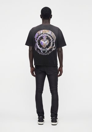 Homme debout de dos, portant un t-shirt noir avec un cœur coloré épineux et un texte stylisé dans le dos, pantalon noir et baskets noires.