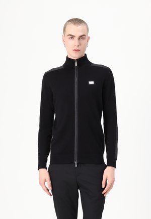 Sudadera negra con cremallera y cuello alto, que cuenta con mangas largas y un discreto parche de logo en el frente. Fabricada con una tela suave y lisa.