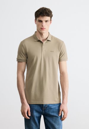 Νέος άνδρας που φοράει μπεζ κοντομάνικο πόλο και μπλε τζιν, στέκεται μπροστά σε ένα απλό, ανοιχτό γκρι φόντο.