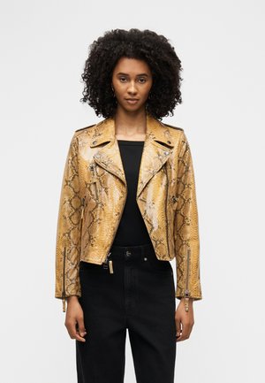 Ženska s kodrastimi lasmi, oblečena v bleščeč motoristični jakni s potiskom kačje kože v barvi bronastega usnja, črno majico in črne hlače, stoji pred enobarvnim ozadjem.