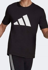 Musta puuvillainen t-paita, jossa on suuri valkoinen kolmonauhalogo edessä. Vakiopituiset pyöreät kaula-aukot ja lyhyet hihat. Istuu tiukasti.