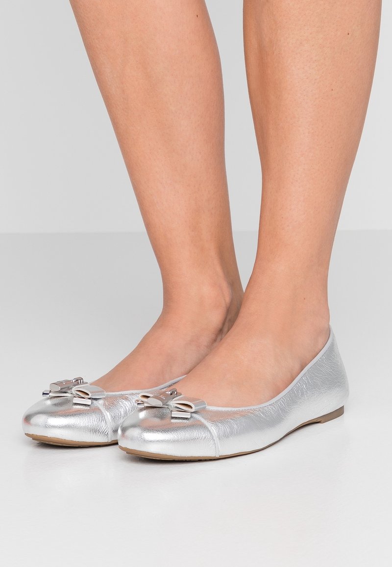 Jambes portant des ballerines métalliques argentées avec des nœuds décoratifs sur le dessus, debout sur un sol gris clair contre un fond gris.