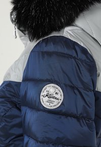 Marineblaue und graue Steppjacke mit schwarzem Fellkragen und rundem weißen Markenlogo auf dem oberen Ärmel.