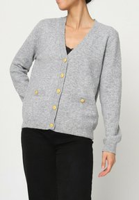 Gilet gris en tissu doux, présentant un col en V, des manches longues, des boutons dorés et une petite poche du côté droit.