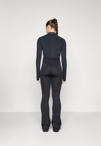 Crna atletska majica dugih rukava i kroja koji se završava iznad struka, uparena s crnim širokim tajicama. Glatka tekstura i prilagođen silueta kroz cijeli outfit.