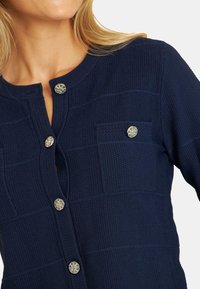 Marineblauer, strukturierten Cardigan mit Rundhalsausschnitt, der markante silberne Knöpfe und zwei Brusttaschen mit einem Karomuster aufweist.