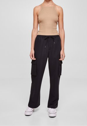 Kobieta w beżowej bezrękawnikowej bluzce z prążkowanego materiału, czarnych spodniach cargo z bocznymi kieszeniami i białych sneakersach na platformie, stojąca na jednolitym tle.