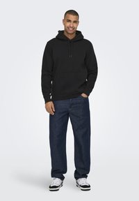 Felpa con cappuccio nera con tasca frontale, abbinata a jeans blu larghi e sneakers bianche e nere. Tessuto dalla texture liscia.