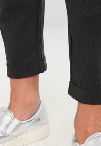 Calças de ganga pretas com bainha, acabamento liso e costura cinza, combinadas com sapatos prateados brilhantes de escorregar e solas brancas.