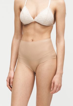 Femme portant un soutien-gorge en dentelle blanche et un sous-vêtement beige taille haute sans couture, posant devant un fond blanc uni.