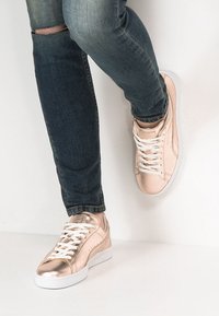 Metalliska roséguld sneakers med vita snören, med en slät yta och rund tå, bärs med mörka rivna jeans. En vanlig vit sula.
