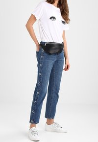 Marsupio nero, t-shirt bianca in cotone con stampa di occhi, jeans di denim blu con dettagli in metallo, e sneakers bianche con decorazioni floreali.