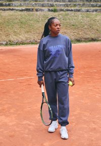 Lila sweatshirt med vit text, matchande joggingsbyxor och vita sneakers. Tennisracket i ena handen och en tennisboll i den andra.